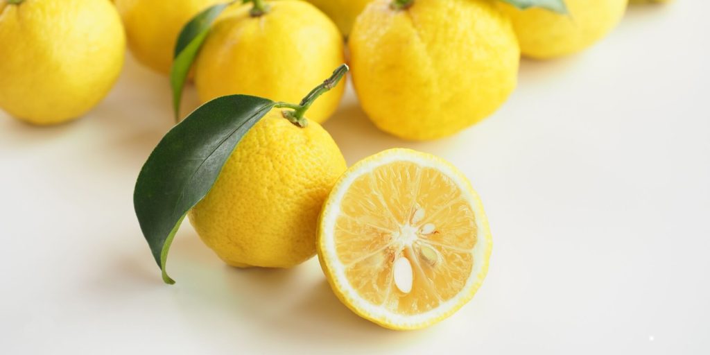 A group of fresh yuzu fruits with bright yellow, slightly rough-textured skin are arranged on a white background. One yuzu is cut in half, revealing its juicy, seed-filled interior. A green leaf is attached to one of the fruits. 