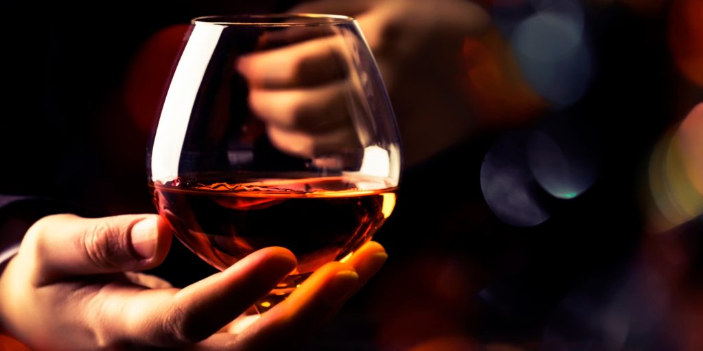 A close-up of a hand elegantly holding a snifter glass filled with amber-colored cognac. The warm glow of the liquid reflects against the glass, while the blurred background features soft, moody lighting with bokeh effects, creating a sophisticated and luxurious atmosphere.