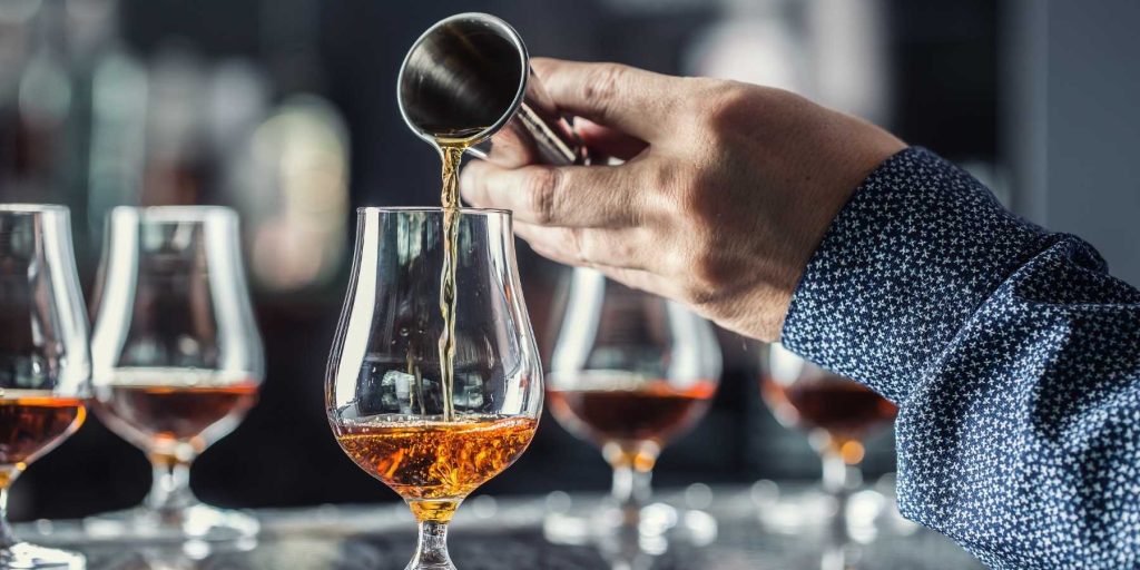 A close-up of a bartender's hand expertly pouring Calvados into a tulip glass during a tasting session, with multiple glasses lined up in the background. Image by weyo. 