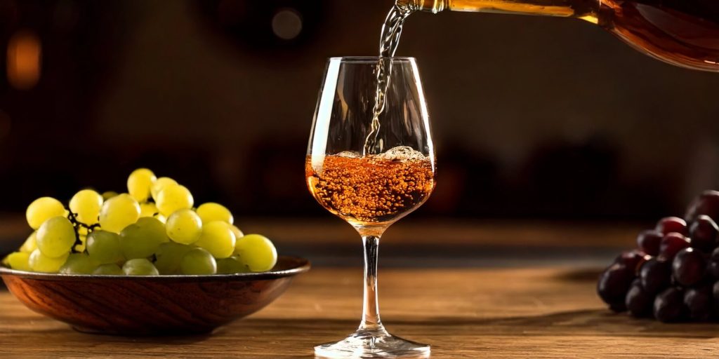 A close-up of a sherry glass being filled with golden liquid, with fresh green and red grapes in the background.