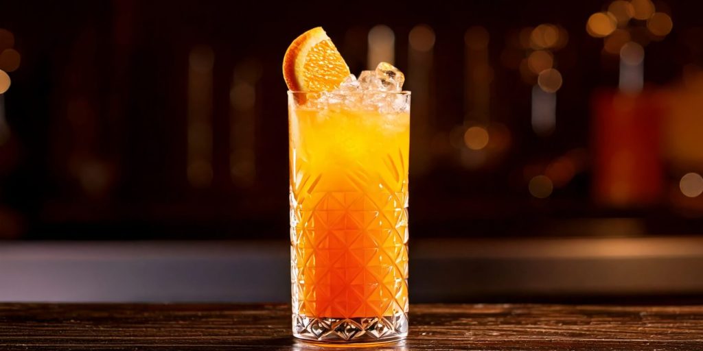 A tall glass of Sherry Cobbler cocktail with crushed ice and an orange garnish, sitting on a bar counter under warm, golden lighting.